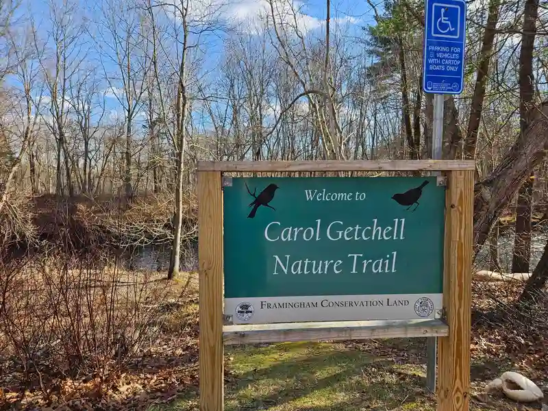 Sign: Welcome to Carol Getchell Nature Trail, Framingham Conservation Land. With bare trees and a little bit of river visible behind
