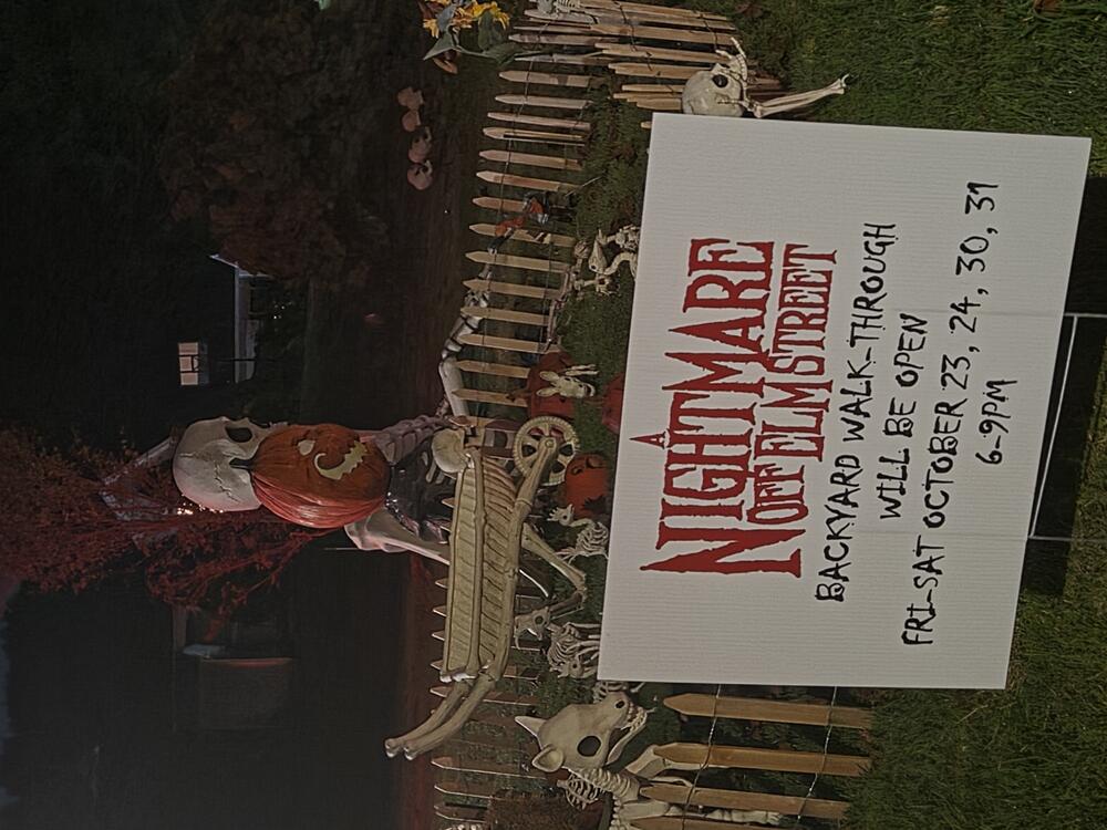 photo of a sign for Nightmare Off Elm Street at night with Halloween decorations behind it. Sign says Backyard walk-through will be open Fri-Sat Oct 23, 24, 30, 31 6-9 PM